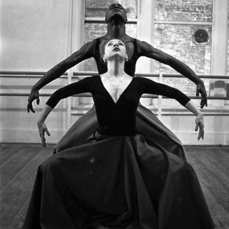 Anna Czajun and Joel Hall Anna Czajun, dressed in a long sleeve leotard and skirt, is standing in front of Joel Hall, who is dressed in a black tank leotard and pants. Both are looking up, knees slightly bent, with their arms outstretched in an upside down "U" position.