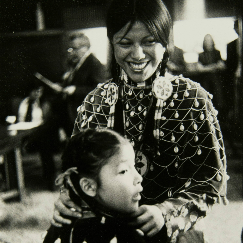 Barbara Bearskin and Kathy White, D'Arcy McNickle in the background during the McNickle Center's founding in 1971 Barbara Bearskin with her hands on Kathy White's shoulders. Both are wearing their powwow regalia. D'Arcy McNickle is in the background of the photo.