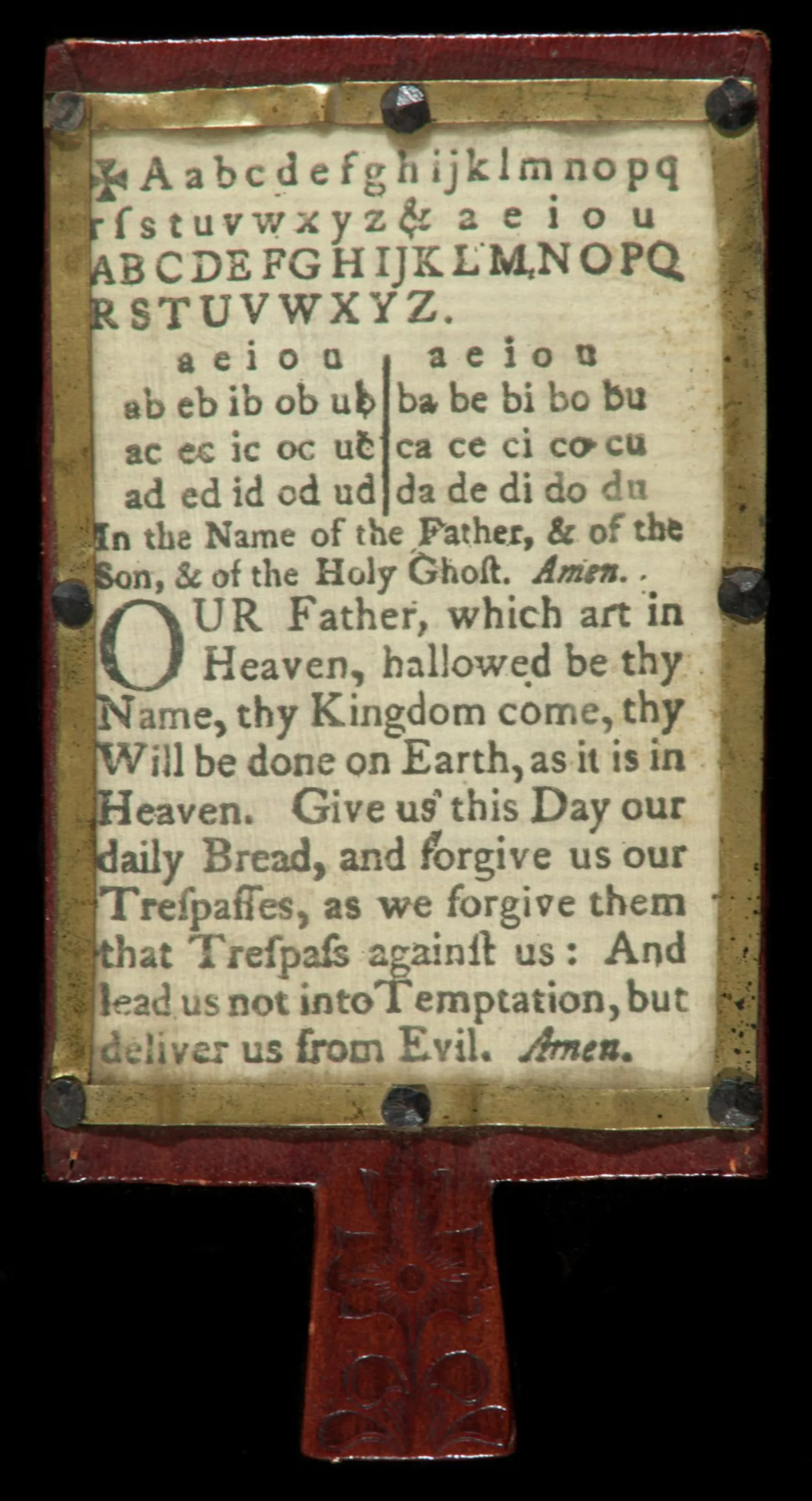 Hornbook with prayer A metal frame is mounted to a wooden board. Within the frame is a printed sheet of paper with the alphabet and the lord’s prayer on it.