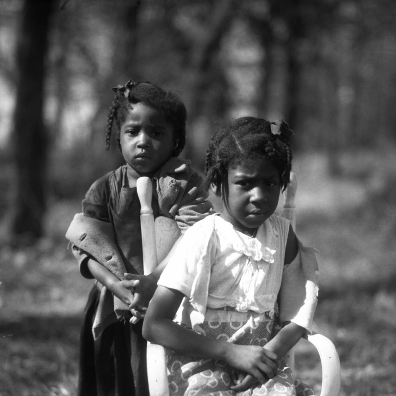 Helen Balfour Morrison photograph Two young girls look into the camera. One of them is seated in a small chair in a field. The other is standing behind the chair.