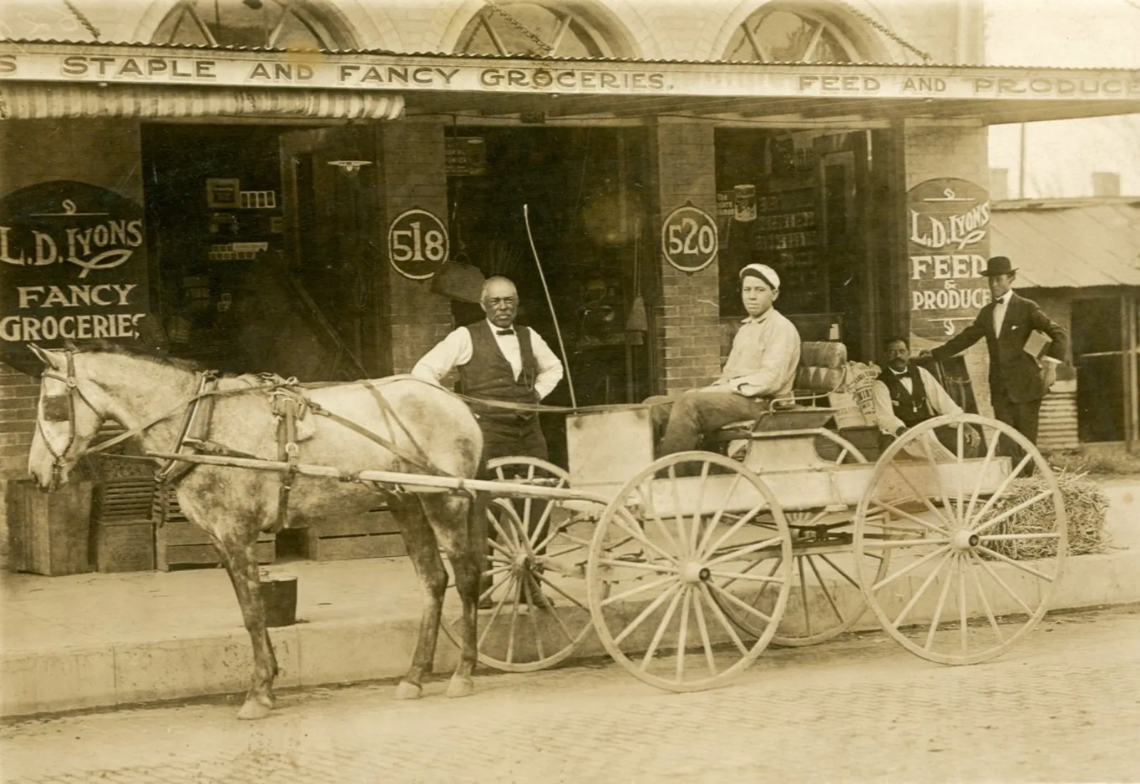 MMS Prescott 009 Lyons Groceries Exterior People pose around a horse and buggy. They're standing in front of a storefront with the words "LD Lyons Fancy Groceries" on the facade.