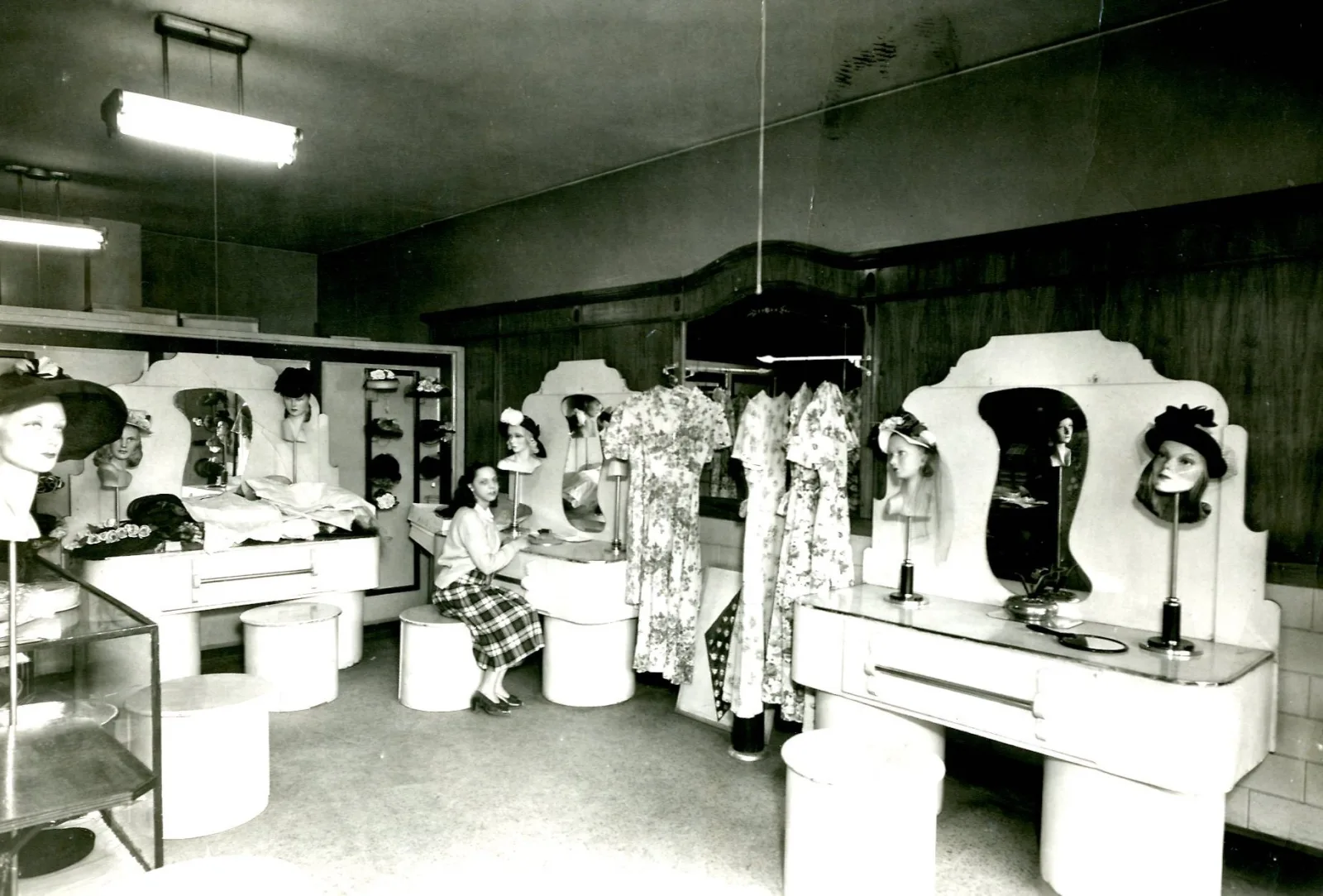 MMS Prescott 038 Lavinia Prescott in Hat Shop A girl sits on a stool surrounded by mannequin heads wearing hats