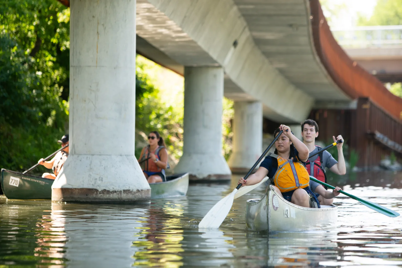 Newberry Canoe 032 NCAIS participants paddle down the Chicago River. Behind them are large concrete poles and highways characteristic of the city's urban landscape.